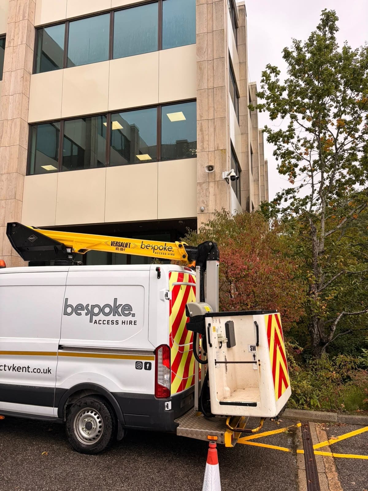 The cherry picker basket lowered behind the Bespoke Access Hire van, below a CCTV dome camera and speaker on the office wall