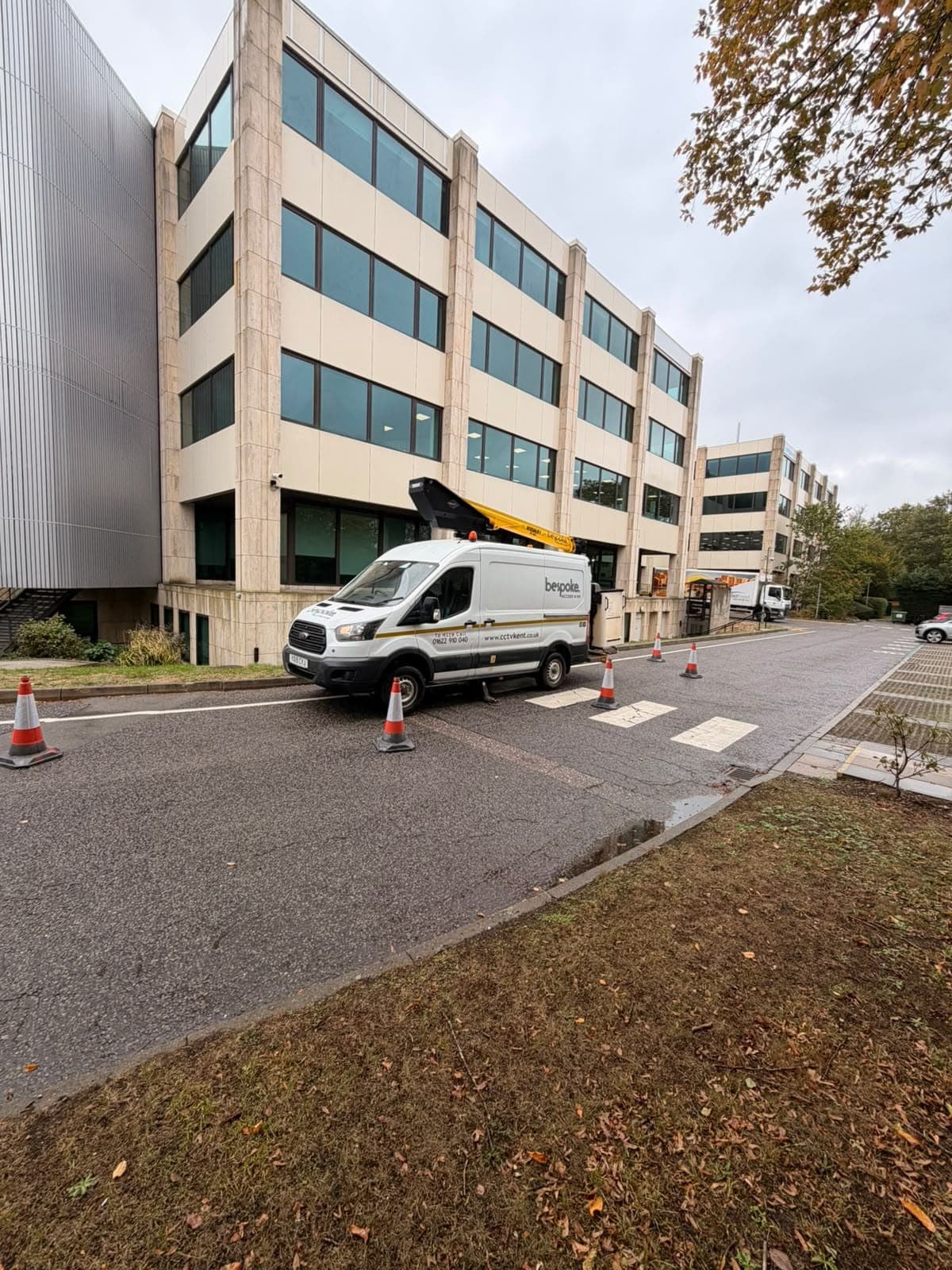 The Bespoke Access Hire van with its boom raised beside an office building on a CCTV maintenance job, the work area coned off on the road